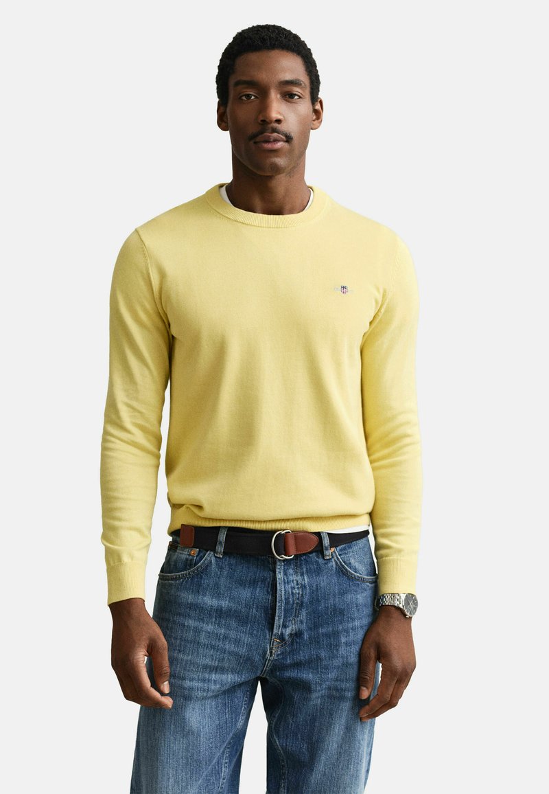 Mann trägt einen gelben Pullover mit langen Ärmeln, blaue Jeans mit schwarzem und braunem Gürtel sowie eine silberne Armbanduhr, steht vor weißem Hintergrund.