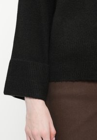 Pull noir en tricot avec une finition côtelée aux poignets et à l'ourlet. Texture douce, présentant une coupe droite et des manches longues, porté sur un tissu brun.