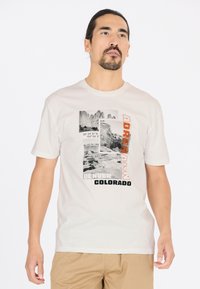 Ljust beige t-shirt med ett grafiskt motiv som har svart och orange text, bergbilder och geografiska koordinater. Kortärmad, rund hals.