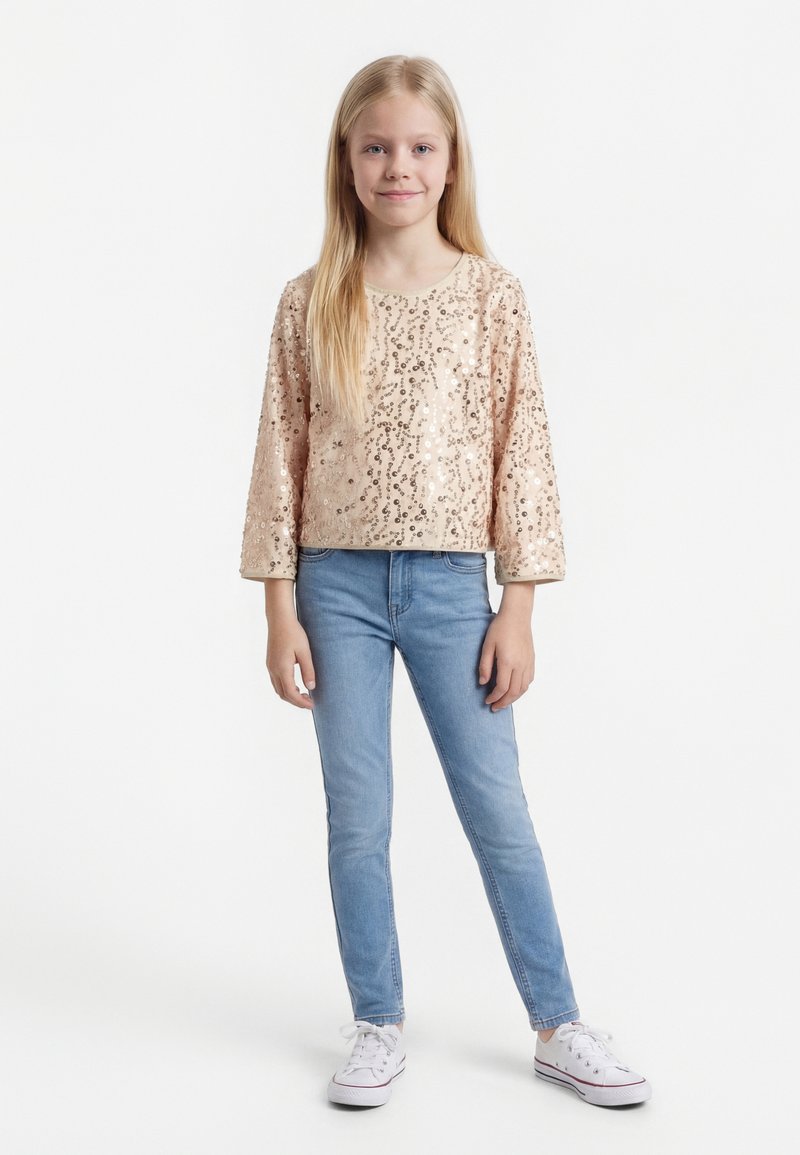 Fille blonde debout portant un top à manches longues beige à paillettes, un jean skinny bleu clair et des baskets blanches sur un fond blanc.