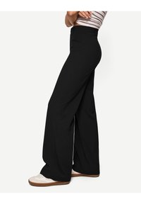 Svarta vida byxor i flexibelt tyg, med hög midja och sidofickor. Bärs med ljusa sportskor.