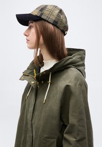 Νεαρή γυναίκα με πράσινο παλτό με κουκούλα και καρό καπέλο που κοιτάζει αριστερά, σε απλό ανοιχτό φόντο.