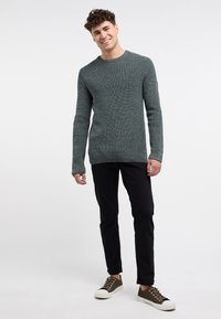 Maglione verde testurizzato con maniche lunghe, collo rotondo, abbinato a pantaloni neri e sneaker marroni. Dettaglio di cucitura evidente sul maglione.