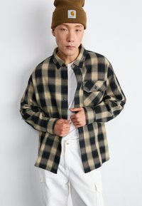 Camisa de franela a cuadros en negro y beige con diseño de botones, un bolsillo en el pecho y botones negros, usada sobre una camiseta blanca y pantalones blancos.