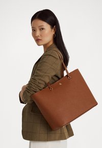 Brown leather tote bag with a smooth texture, featuring matching handles and a gold logo on the front. Structured rectangular shape.