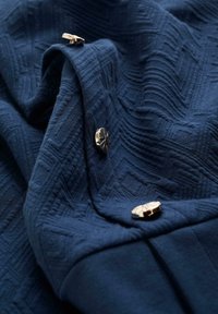 Marinblått textil med ett geometriskt mönster, prytt med metallknappar. Tyget verkar mjukt och hänger smidigt.