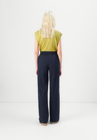 Femme aux longs cheveux blonds se tient face à un mur blanc, portant un haut vert sans manches et un pantalon large taille haute bleu marine.