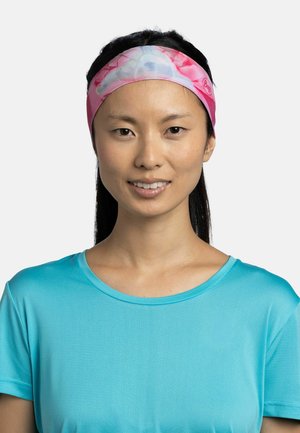 Mujer con cabello largo y oscuro que lleva una diadema rosa y azul con estampado y una camiseta deportiva azul claro, sonriendo sobre un fondo blanco.