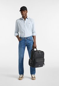 Mochila negra con bolsillos frontales, sostenida con una mano. Una camisa de rayas azul claro, jeans azules y zapatillas beige completan el atuendo.