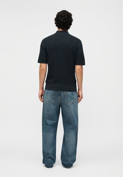 Άνδρας με σγουρά μαλλιά στέκεται γυρισμένος προς τα πίσω, φοράει μαύρο κοντομάνικο πουκάμισο και άνετο μπλε τζιν πάνω σε απλό φόντο.