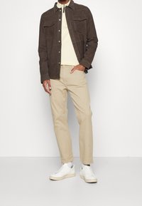 Brązowa zamszowa kurtka z dwoma przednimi kieszeniami, beżowa koszulka polo, jasnobeżowe spodnie oraz białe trampki na neutralnym tle.