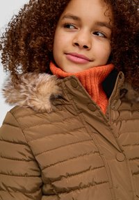 Braune puffige Jacke mit einem mit Fell gefütterten Kapuzen, die über horizontale Steppnähte und Knopfverschlüsse verfügt, kombiniert mit einem orangefarbenen, gerippten Rollkragenpullover.