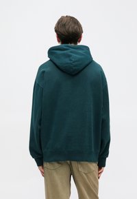 Haut à capuche surdimensionné en teal avec un cordon de serrage, ourlet et poignets côtelés. La vue arrière montre une texture de tissu lisse et une coupe décontractée. Associé à un pantalon beige.