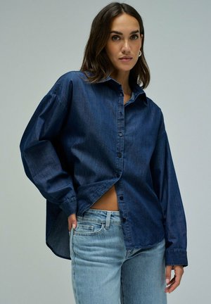 Femme portant une chemise en denim bleu foncé oversized et un jean bleu clair, debout devant un fond uni, main dans la poche, souriant légèrement.