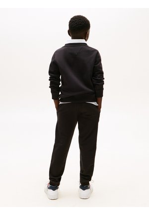 Barn stående med ryggen til, iført sort sweatshirt, sorte bukser og hvide sneakers med blå accenter på en ensfarvet hvid baggrund.