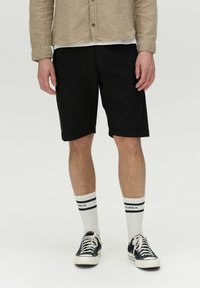 Schwarze Baumwollshorts im geraden Schnitt, kombiniert mit weißen knielangen Socken mit schwarzen Streifen und schwarzen Turnschuhen.