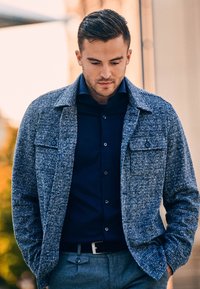 Op maat gemaakte blauwgrijze gestructureerde jas met grote borstzakken, gedragen over een donkerblauw overhemd met knopen, aangevuld met grijze nette broek.