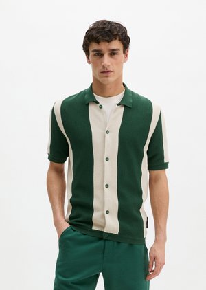 Fiatal férfi zöld-fehér függőleges csíkos rövid ujjú gombos ingben és zöld nadrágban, egyik kezével a zsebében áll.