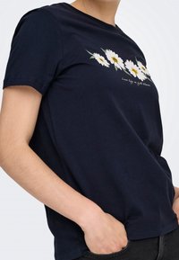 Personne portant un t-shirt noir avec des imprimés de marguerites blanches et le texte "Live life in full bloom" sur la poitrine, main dans la poche.