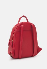 Sac à dos rouge de forme arrondie, fabriqué en matériau synthétique lisse. Il possède deux bretelles réglables, une poignée supérieure et des éléments métalliques dorés.