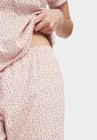 Una mano levantando una blusa rosa con lunares que revela unos pantalones elásticos a juego contra un fondo neutro.