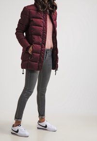 Bordowa kurtka puchowa zapinana na zamek, w połączeniu z jasnoróżowym swetrem, szarymi jeansami o wąskim kroju oraz białymi sneakersami Nike z czarnymi akcentami.