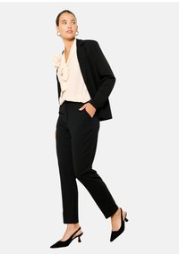 Črna prilagojena blazer jakna preko bež bluze z volančki, v kombinaciji s črnimi oprijetimi hlačami in črnimi slingback petami. Preprost dizajn in čiste linije.
