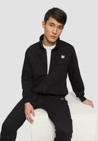 Schwarze Zip-up Sweatjacke aus weichem Stoff, mit einem kleinen goldenen Logo auf der Brust, kombiniert mit passenden schwarzen Hosen und einem orangefarbenen Akzent.