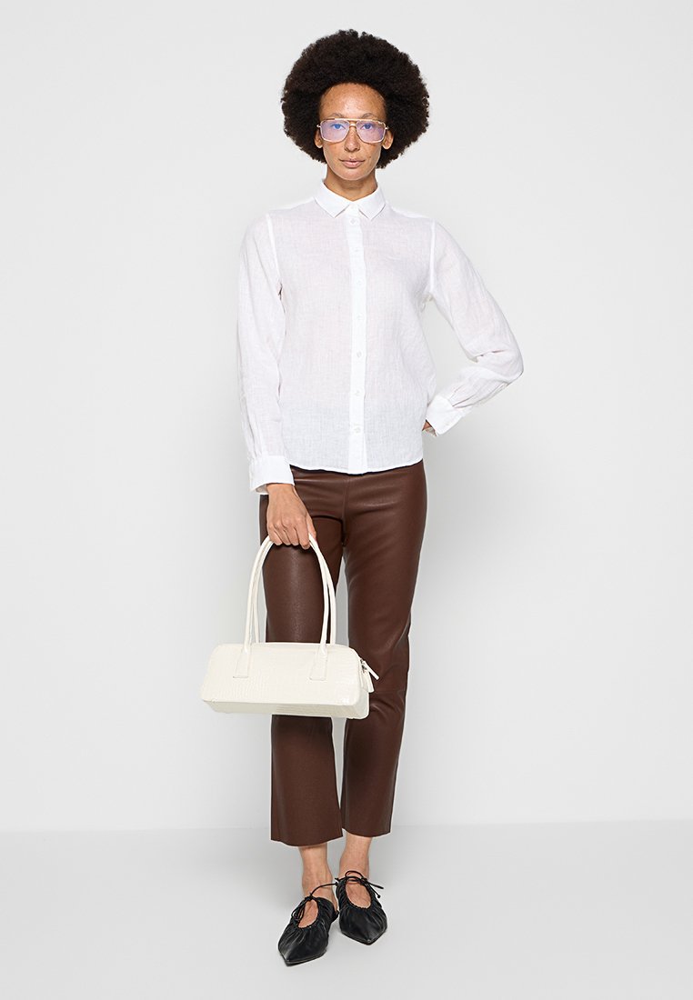 Biała koszula zapinana na guziki, brązowe skórzane spodnie, czarne buty z witkami i mała biała torebka o fakturze. Przezroczyste okulary i kręcone włosy.