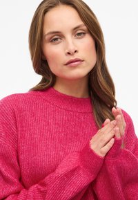 Jasnoróżowy prążkowany sweter z wysokim kołnierzem i długimi rękawami. Posiada otwory na palce przy mankietach. Miękka tekstura z delikatnym połyskiem.