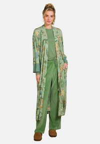 Grüner, blumiger langer Kimono mit gestreiftem Bündchen, übereinander getragen mit einem passenden grünen Shirt und einer Hose. Weicher Stoff mit lockerer Passform. Braune Schuhe.