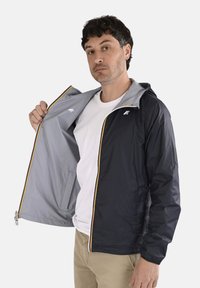 Homme portant une veste légère noire ouverte pour montrer la doublure intérieure grise, associée à un t-shirt blanc et un pantalon beige, sur un fond uni.