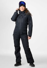 Zwart geïsoleerd sneeuwpak met meerdere ritszakken, hoge kraag en verstelbare manchetten. Draagt een zwarte beanie en skibril.