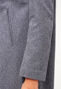 Abrigo de lana gris con una textura suave, que cuenta con un solo bolsillo lateral y un corte de manga recta. El material parece cálido y grueso.