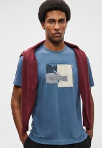 T-shirt de algodão azul com um design gráfico em várias tonalidades, com um suéter bordô pousado sobre os ombros. Mangas curtas, decote redondo.