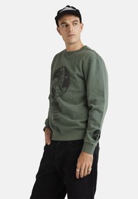 Green sweatshirt with a round graphic and text print. Ribbed cuffs and hem. Model wears black jeans and a black cap.