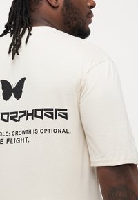Cream short-sleeve t-shirt featuring a black butterfly graphic and bold text on the back: "MORPHOSIS; GROWTH IS OPTIONAL. TAKE FLIGHT."