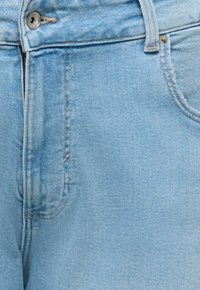 Svetlomodré rifľové nohavice zblízka zobrazujúce predný gombík, zips, očko na opasok a detaily šitia vrecka.