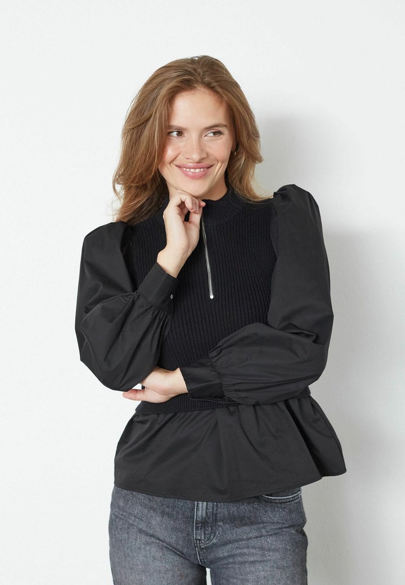 Črn peplum top z rebrastim bodicem, visokim ovratnikom in zadrgo; ima obsežne balonske rokave iz gladkega materiala.