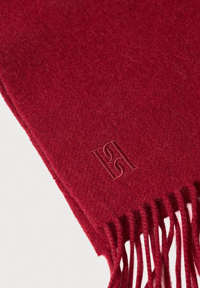 Červená vlněná šála s roztřepenými konci a jemným vyraženým logem. Textura působí měkce a teple, vhodná pro chladné počasí.