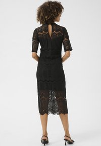 Vestido negro de encaje con un patrón floral, mangas cortas y un detalle en forma de cerradura en la espalda; el dobladillo inferior transparente revela la piel, combinado con tacones negros.