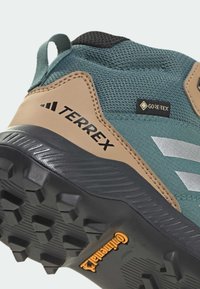 Blågrønn og beige tursko har et GORE-TEX-merke, teksturert mesh-overdel, gummisåle med fremtredende mønster og en svart bunn.