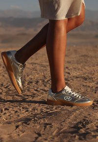 Silver- och guldskor med en slät metallisk finish, ovandel av läder och gummisula. Har snörning och en framträdande logotyp på sidan.