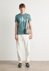 Žalios medvilnės marškinėliai su balta Calvin Klein Jeans logotipu, derinami su kreminėmis kelnėmis ir daugiaspalviais sportbačiais, papildytas šviesiai rudos spalvos petine rankine.