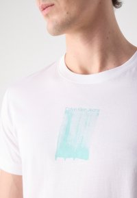 Biała bawełniana koszulka z grafiką w turkusowych pociągnięciach pędzla oraz napisem "Calvin Klein Jeans" w nowoczesnej czcionce. Krótkie rękawy i okrągły dekolt.