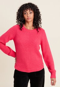 Rosa Strickpullover mit Zopfmuster, Rundhalsausschnitt, gerippten Bündchen und Knöpfen an den Ärmeln. Kombiniert mit schwarzen Cordhosen.