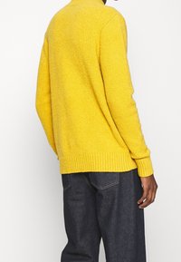 Gelber Strickpullover mit texturiertem Finish, gerippten Bündchen und Saum. Über dunklen Jeans getragen, was eine lässige Passform zeigt.