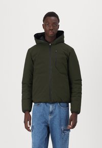 Veste rembourrée à capuche en tissu vert foncé avec une fermeture éclair à l'avant, des poignets ajustables et une petite poche sur la poitrine. Portée avec un jean cargo bleu clair.