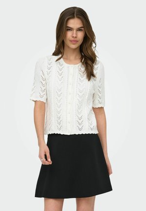 Femme portant un haut en maille blanc à manches courtes, boutonné avec un motif chevron, associé à une jupe noire taille A, se tenant devant un fond uni.