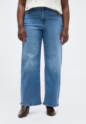 Person iført blå vidde jeans, hvid top med små orange print og brune ankelstøvler med hæl, stående på en neutral baggrund.